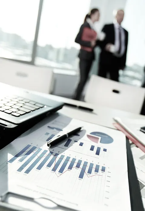 Close-up of a desk with a laptop, printed charts, and pens, while two businesspeople stand and talk blurred in the background.