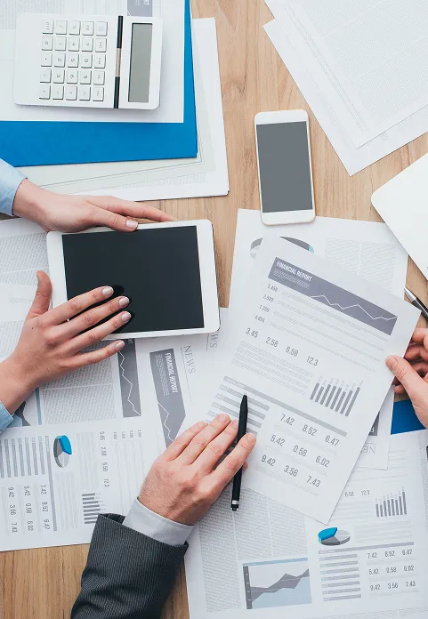 Top-down view of a desk with a tablet, papers with charts, and a laptop; hands of people are working on documents.